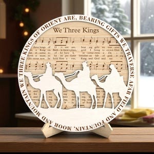 May include: A circular wooden Christmas ornament with the words "We Three Kings" and a silhouette of the three wise men on camels. The background features sheet music. The ornament is displayed on a wooden stand.