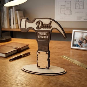 May include: A wooden hammer-shaped desk ornament with the word "Dad" and the phrase "You Built My World" engraved on it. The ornament is made of light and dark wood and sits on a wooden base. A thoughtful gift.
