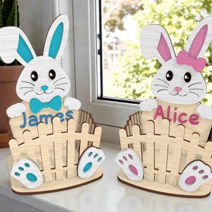 Puede incluir: Dos decoraciones de conejos de Pascua de madera. Un conejo tiene detalles en azul y el nombre "James" en azul. El otro conejo tiene detalles en rosa y el nombre "Alice" en rosa. Ambos conejos sostienen una valla de madera.