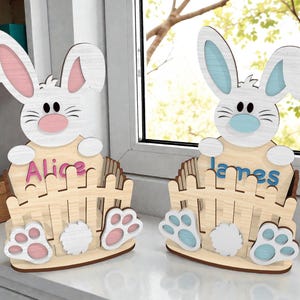 May include: Two wooden Easter bunny ornaments. One bunny has pink ears and nose, the other has blue. Each bunny holds a name sign, and is set in front of a wooden fence with paw prints.