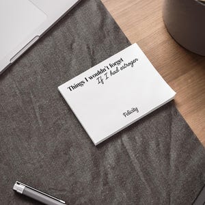 Pode incluir: Um bloco de notas branco com o texto "Things I wouldn't forget If I had estrogen" e o nome "Felicity" em preto. O bloco de notas está sobre uma superfície cinza escuro com um laptop e uma caneta visíveis.