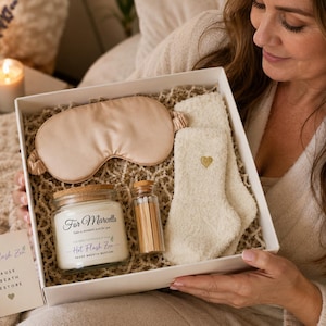 May include: A gift box containing a champagne-colored satin sleep mask, a pair of cream-colored fuzzy socks with a small gold heart embroidery, a glass candle jar with a cork lid labeled For Marcella Hot Flash Zen, and a small glass vial of matches.