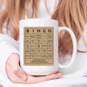 White ceramic mug featuring a vintage-style menopause bingo card design with common symptoms like brain fog, hot flashes, and mood swings. Woman holding mug shown in 15oz size.  Back personalized in black.