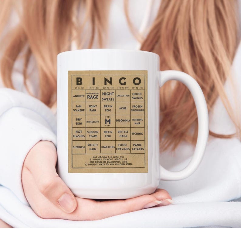 White ceramic mug featuring a vintage-style menopause bingo card design with common symptoms like brain fog, hot flashes, and mood swings. Woman holding mug shown in 15oz size.  Back personalized in black.