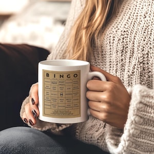 Woman in a beige sweater holding a white ceramic mug featuring a vintage-style menopause bingo card design with common symptoms like brain fog, hot flashes, and mood swings. Mug shown in 11oz and 15oz sizes.  Back personalized in black.