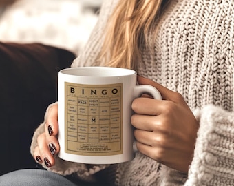 Puede incluir: Una taza de cerámica blanca con un gráfico rectangular de color tostado impreso en el frente. El gráfico es un cartón de bingo titulado BINGO con varias etiquetas de texto en cada casilla, incluyendo anxiety, rage, night sweats y brain fog.