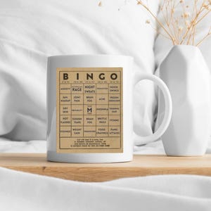 White ceramic mug featuring a vintage-style menopause bingo card design with common symptoms like brain fog, hot flashes, and mood swings. Mug shown on a wooden tray with dried flowers behind it.