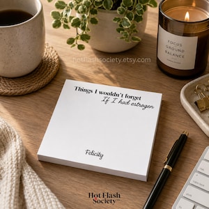 May include: A square white notepad featuring the black printed text 'Things I wouldn't forget If I had estrogen' at the top and the name 'Felicity' at the bottom, positioned on a wooden surface with a black pen and other desk items.