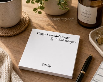May include: A square white notepad featuring the black printed text 'Things I wouldn't forget If I had estrogen' at the top and the name 'Felicity' at the bottom, positioned on a wooden surface with a black pen and other desk items.