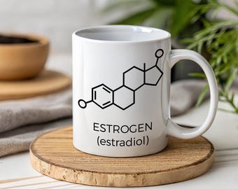 May include: A white ceramic mug featuring a black chemical structure diagram of estrogen and the text "ESTROGEN (estradiol)" printed on the front. The mug has a standard cylindrical shape with a curved handle on the right side.