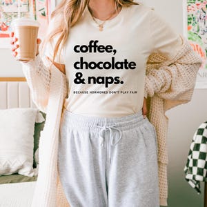 Könnte beinhalten: Ein cremefarbenes Kurzarm-T-Shirt mit dem schwarzen Text 'coffee, chocolate & naps.' und 'BECAUSE HORMONES DON'T PLAY FAIR' auf der Vorderseite, getragen mit einer beigen Strickjacke und einer hellgrauen Jogginghose mit Kordelzug.
