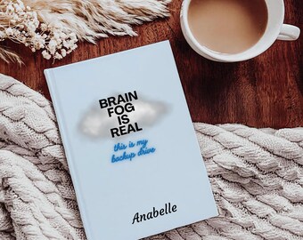 Puede incluir: Un libro azul claro con el texto "BRAIN FOG IS REAL" y "this is my backup drive". El libro está sobre una superficie de madera junto a una taza de café y una manta de punto blanca.