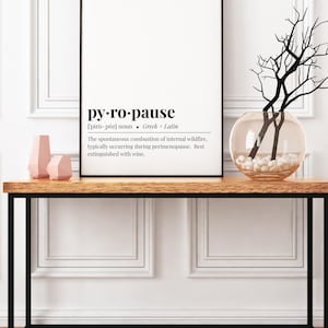 May include: A framed print with the word "py-ro-pause" and its definition. The print is on a wooden table with a black metal frame, alongside decorative vases and a glass vase with branches.