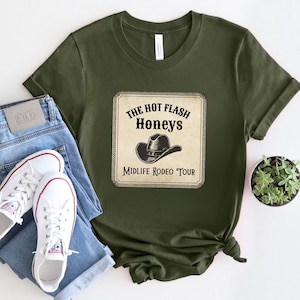 May include: Olive green t-shirt featuring a graphic with the text "The Hot Flash Honeys Midlife Rodeo Tour" and a cowboy hat illustration. The shirt is paired with blue jeans and white sneakers.