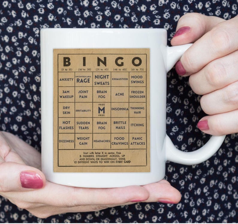 White ceramic mug featuring a vintage-style menopause bingo card design with common symptoms like brain fog, hot flashes, and mood swings. Woman holding 11oz mug infront of a navy blue print blouse.  Back personalized in black.