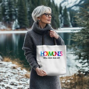 Può includere: Borsa tote bianca con manici neri. La borsa presenta la parola "HOMINES" nei colori dell'arcobaleno, con il testo "WILL KICK YOUR ASS" sotto. Una persona con un maglione grigio tiene la borsa, con un lago e alberi sullo sfondo.
