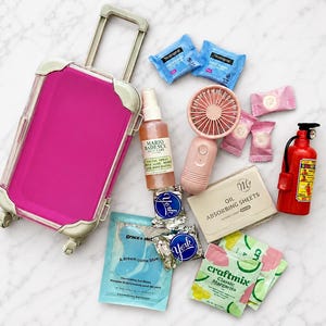 Op de afbeelding: Een felroze rolkoffer met zilveren accenten, vergezeld door een verzameling reisartikelen, waaronder een Mario Badescu gezichtsspray, een roze handventilator, olie-absorberende velletjes, York pepermuntjes en een rode brandblusser.