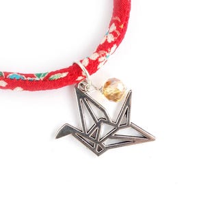 Può includere: Un braccialetto in tessuto rosso con un motivo floreale bianco, verde e blu. Un ciondolo in metallo argentato a forma di gru origami pende dal cinturino, accompagnato da una piccola perla sfaccettata traslucida color ambra chiaro.