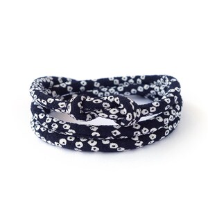 May include: A dark navy blue fabric hair tie featuring a repeating white circular pattern. The accessory is constructed from a continuous band of material that is knotted at the top, creating a layered, circular shape with a soft, flexible texture.