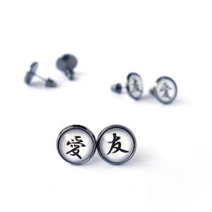 May include: Round stud earrings with a black border and a white background, each displaying a black Japanese character. Other earrings and backs are visible in the background. The earrings are small.