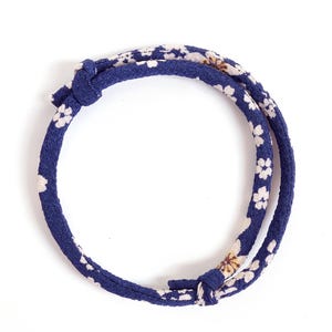 May include: A circular fabric bracelet featuring a deep blue base color with a repeating pattern of small white and light brown floral blossoms. The bracelet is constructed from a continuous cord of fabric that is secured with two visible knots.