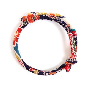 May include: A circular fabric bracelet featuring a vibrant floral pattern in shades of navy blue, red, orange, and white. The textile band is tied with two distinct knots, creating a continuous loop design with a textured, woven appearance.