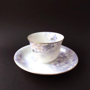 May include: A white teacup and saucer set with a floral design. The cup and saucer have a lavender and white gradient with pink and white cherry blossom accents. The rims are trimmed with gold.