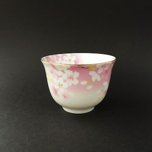 May include: A ceramic teacup with a cherry blossom design. The cup has a gold rim and a rounded shape. The design features white flowers with yellow centers against a pink gradient. The teacup is white at the base and measures approximately 8 cm in height.