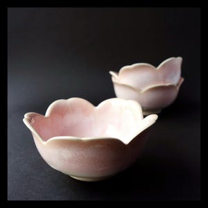 Op de afbeelding: Twee keramische kommen met een lichtroze glazuur en een geschulpte, bloemvormige rand. De kommen hebben een gladde, glanzende afwerking met subtiele kleurvariaties, variërend van een zachte witte basis tot een delicate roze tint langs de randen.