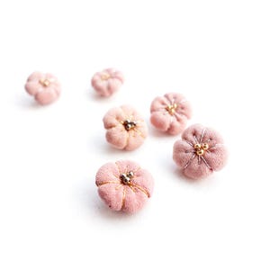 May include: Six small, round, fabric-covered buttons in a soft dusty pink color. Each button is shaped like a five-petaled flower with visible stitching radiating from the center, which is accented with three small, metallic gold-toned beads.