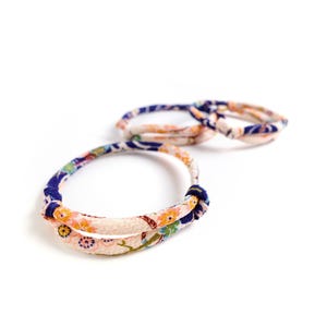 Könnte beinhalten: Drei Stoffarmbänder mit floralen Mustern in Rosa-, Orange-, Blau- und Grüntönen. Die Armbänder sind geknotet und scheinen aus einem weichen, strukturierten Material zu bestehen. Der Hintergrund ist schlicht weiß.