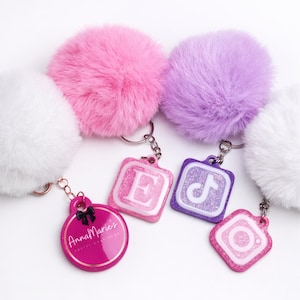 May include: Keychains with fluffy pom-poms in white, pink, and purple. The keychains have pink, purple, and white glittery charms with the letter "E", a TikTok logo, an Instagram logo, and a round charm with the text "Anna Marie's Pretty Creations".