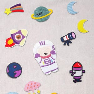 May include: A collection of embroidered space-themed patches. Designs include a shooting star, planets with rings, crescent moons, a rocket ship, a telescope, a bear astronaut, and a cloud with stars and moons. The patches feature bright colors.