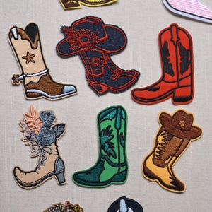May include: A collection of embroidered patches featuring various boot and shoe designs. Included are cowboy boots, a high heel, and boots with floral and star details. The patches are in a variety of colors, including pink, yellow, red, and green.