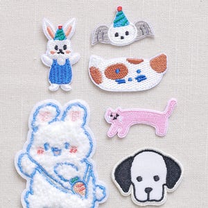 May include: A collection of embroidered patches featuring various animal designs. Includes a white bunny in blue overalls, a pink cat, a white dog with black ears, and other whimsical creatures. Each patch has a colorful, cartoonish style.