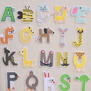 Puede incluir: Letras del alfabeto bordadas, cada una diseñada como un animal diferente. Las letras son de varios colores, incluyendo verde, naranja, amarillo y azul. Las letras están cosidas sobre una tela beige claro.