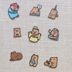 Peut inclure: Collection de neuf écussons brodés représentant des capybaras de dessins animés dans diverses poses et tenues. Les écussons incluent des capybaras portant des robes, des chapeaux et tenant des objets comme des poissons et un ordinateur. Les écussons sont dans des tons de marron, beige et bleu.