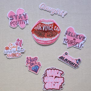 Pode incluir: Uma coleção de patches bordados em tons de rosa e branco. Os patches apresentam várias frases motivacionais como "Stay Positive", "Believe in Yourself" e "Make Today Great". Outros patches incluem um tema de cowgirl e um diamante.