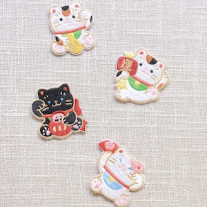 以下が含まれることがあります： 白、黒、ピンクの日本の招き猫、またはラッキーキャットの刺繍パッチ。 金貨や幸運の漢字などの詳細が特徴です。 衣類やアクセサリーに加えるのに最適です。