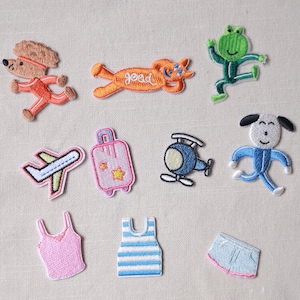 Puede incluir: Colección de parches bordados con diversos diseños. Incluye un perro, un gato, una rana, un avión, una maleta, un helicóptero y un personaje parecido a un perro. También, una camiseta sin mangas rosa, una camiseta sin mangas a rayas azules y blancas, pantalones cortos vaqueros y pantalones cortos azules.