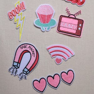 May include: A collection of embroidered patches in various shapes and colors. Designs include a magnet with "Babe Magnet" text, a paper airplane with "Keep going" text, the word "Hello", a lightning bolt with "BOOM!" text, a pink cupcake, a pink TV with "CRAZY" text, a wifi symbol, and pink hearts.