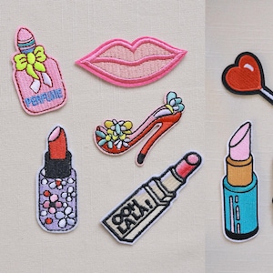 Może przedstawiać: Kolekcja haftowanych naszywek o tematyce urody i mody. Zawiera butelkę perfum, usta, szpilkę, szminki, lizaka w kształcie serca, wskaźniki naładowania baterii oraz aparat fotograficzny z uśmiechniętą twarzą. Dołączona jest również naszywka z napisem "Ooh La La!".
