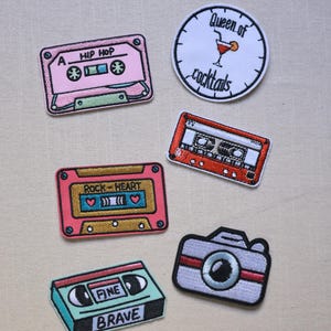 Könnte beinhalten: Eine Sammlung von aufgestickten Aufnähern mit Retro-Designs. Enthält ein rosa Kassettenband mit dem Text "HIP HOP", einen runden Aufnäher mit einem Cocktail und dem Text "Queen of cocktails" und einen Kamera-Aufnäher.