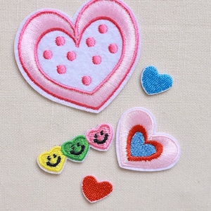 May include: A collection of embroidered heart patches in various sizes and colors. The largest heart is pink with a white center and pink polka dots. Other hearts are blue, red, and multi-colored with smiley faces.