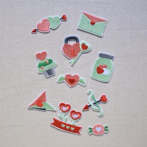 Puede incluir: Una colección de parches bordados con temas de San Valentín. Los diseños incluyen corazones, un sobre, una magdalena, una cerradura y llave, un tarro, un arco y flecha, flores y caramelos. Los parches son rojos, verdes y blancos.