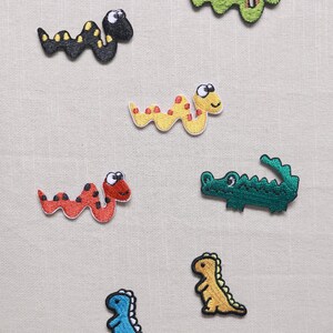 May include: A selection of embroidered patches depicting cartoon animals. The patches include a black snake with yellow spots, a green snake, a yellow snake with red spots, a red snake, a green alligator, a blue dinosaur, and a yellow dinosaur.