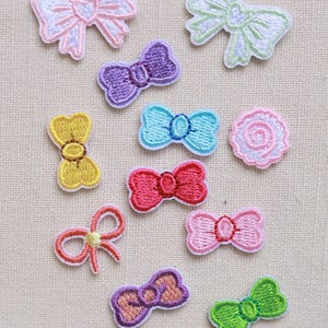 Puede incluir: Colección de parches bordados con forma de lazo en varios colores, incluyendo rosa, morado, amarillo, azul, rojo y verde. Cada parche presenta un diseño detallado de lazo con contorno blanco, adecuado para manualidades y adornos.