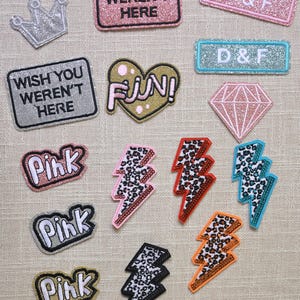 Puede incluir: Una colección de parches decorativos termoadhesivos en varias formas y colores. Los diseños incluyen rayos, corazones, diamantes, coronas y parches rectangulares con textos como "Pink", "Fun!" y "Wish You Weren't Here".