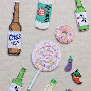 Puede incluir: Parches bordados de varios alimentos y bebidas, incluyendo botellas de cerveza y soju, una piruleta, una rosquilla y verduras. Los parches son coloridos y detallados, con texto en algunas botellas.