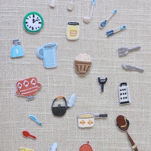 May include: A collection of embroidered kitchen-themed patches. Includes a clock, jar, measuring cup, cupcake, teapot, pan, spoon, and various utensils. Patches are in various colors, including blue, red, and brown, and are arranged on a neutral background.